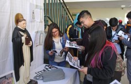 Imagen de Santo Tomás en una mesa, mientras tres jóvenes leen historieta alusiva.